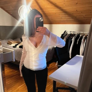 Vit omlottopp med spetsdetaljer - Säljer en elegant vit omlottopp med spetsdetaljer vid halsringningen. Toppen har långa ärmar och en v-ringad design som ger en stilren look. Perfekt för att klä upp eller ner beroende tillfälle! Storlek M men kan va lite större också! 