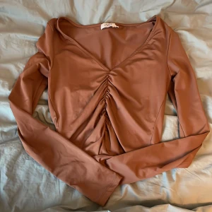 Brun långärmad topp från NA-KD - Säljer en snygg brun långärmad topp från NA-KD med v-ringning och rynkdetalj framtill. Perfekt för en stilren look.