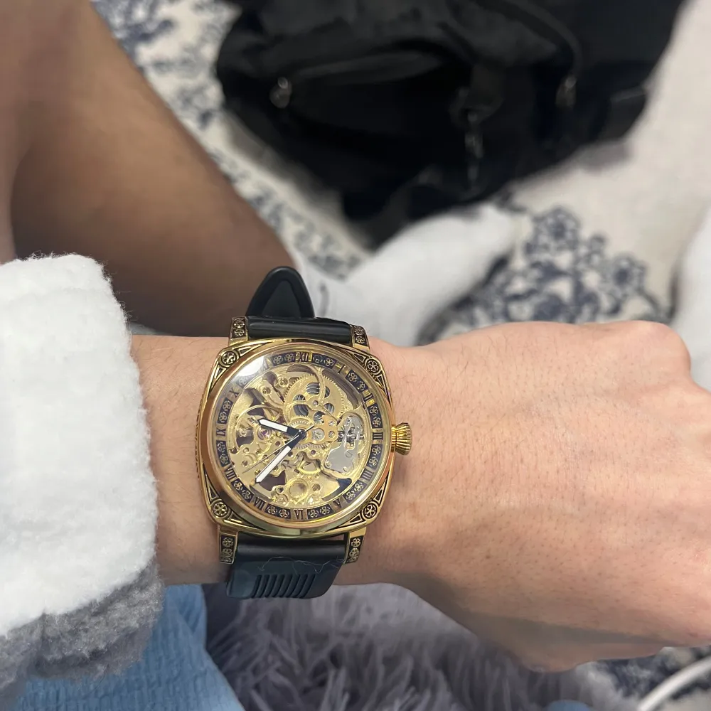 Snygg guldfärgad klocka med synlig urtavla och svart armband. Klockan har en detaljerad design med synliga kugghjul och en robust känsla. Perfekt för den som gillar en iögonfallande accessoar.. Asusteet.