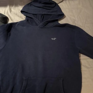 Mörkblå hoodie från Hollister - Säljer en stilren mörkblå hoodie från Hollister med en liten logga på bröstet. Tröjan har en klassisk känguruficka och en bekväm huva. Perfekt för en avslappnad stil. Nypris cirka 450kr. Säljer då den är för liten. Skriv vid minsta fundering. 