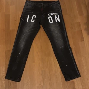 Svarta jeansbyxor från Dsquared2 - Snygga svarta jeansbyxor från Dsquared2 med vit text 'ICON' på framsidan. Byxorna har en cool, sliten look med små färgstänk och en kedjedetalj baktill. Perfekta för en trendig stil. Kan prata om pris samma med alla andra kläder som finns i min profil 