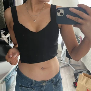 Svart croppad topp från H&M - Säljer en svart croppad topp från H&M med breda axelband och omlott-design. Perfekt för en stilren look. Toppen är ärmlös och har en tight passform. Dragkedja på sidan