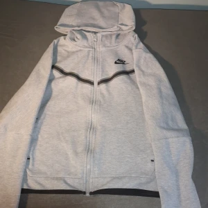 Nike tech grå - Säljer Nike tech med dragkedja och svart logotyp på bröstet. Tröjan har en huva och svarta detaljer längs ärmarna. Perfekt för en sportig look!