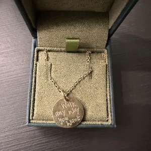 Halsband från Maria Nilsdotter - Säljer ett halsband från Maria Nilsdotter med en rund berlock. Ena sidan har texten 'Walk any way you want' och den andra sidan har ett motiv av fotavtryck. Halsbandet är i guldpläterat silver och kedjan är 45 cm lång. Knappt använd så i ny skick. 