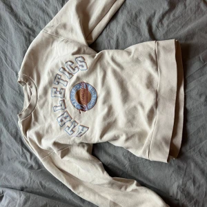 Beige sweatshirt med tryck - Säljer en beige sweatshirt med texten 'Athletics' och ett runt tryck i mitten. Tröjan har långa ärmar och ribbade muddar vid ärmslut och nederkant. Perfekt för en avslappnad stil.