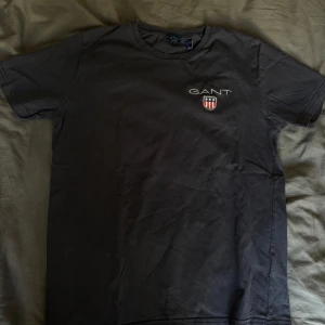 Marinblå t-shirt från GANT - Säljer en stilren marinblå t-shirt från GANT med broderad logga och emblem på bröstet. Perfekt för en casual look. Passar bra till jeans eller shorts.