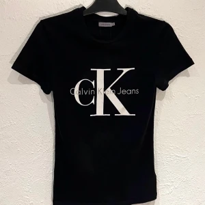 Calvin Klein Jeans - Dam T-shirt - Strl XS - Snygg svart t-shirt från Calvin Klein Jeans med stort vitt CK-logotyptryck på framsidan. Perfekt för en stilren och modern look. Tillverkad i mjukt material för extra komfort. 100% bomull
