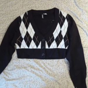 Svart och vit kofta från H&M - Snygg svart och vit kofta från H&M med ett klassiskt argylemönster. Den är croppad och har en v-ringad design med knappar framtill. Perfekt för att lägga till en stilren touch till din outfit. Skriv gärna om du vill ha mer information om koftan 😊