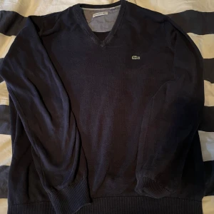 Svart tröja från Lacoste - Säljer en stilren svart tröja från Lacoste med v-ringning och det klassiska krokodilmärket på bröstet. Tröjan har långa ärmar och ribbade muddar vid ärmslut och nederkant. Perfekt för en avslappnad men snygg look.
