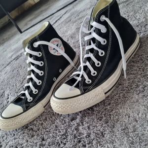 Svarta Converse All Star sneakers - Klassiska svarta Converse All Star sneakers i hög modell med vita snören och sula. Perfekta för en avslappnad stil. Tillverkade i textil med den ikoniska loggan på sidan. Storlek 38