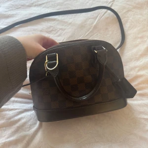 Brun rutig axelväska - Snygg brun rutig axelväska med guldfärgade detaljer och dragkedja. Väskan har en justerbar axelrem och handtag för enkel bärning. Insidan är klädd i rött tyg och det finns en medföljande låsdetalj som gått av men det går att fixa 