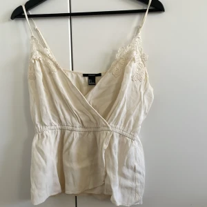 Beige linne med spetsdetaljer - Säljer ett sött beige linne med spetsdetaljer vid axelbanden. Linnet har en omlott-design och tunna axelband, perfekt för varma dagar. Passar bra till både jeans och kjol.