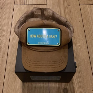 Brun keps från John Hatter & Co - Snygg brun keps med nätpaneler och en blå skylt framtill med texten 'How about a hug?'. Perfekt för en avslappnad stil.