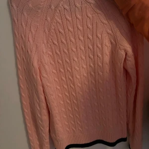 Rosa stickad tröja från Calvin Klein - Säljer en snygg rosa stickad tröja från Calvin Klein med kabelstickat mönster. Tröjan har långa ärmar och en svart rand vid nederkanten. Perfekt för en stilren look!