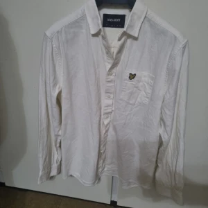 Vit skjorta från Lyle & Scott nyskick - Vit långärmad skjorta från Lyle & Scott med klassisk krage och knäppning framtill. Bröstficka med den ikoniska gula fågel-loggan. Skjortan är i bomull och har en avslappnad passform, perfekt för en clean och stilren look.