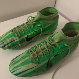 Nike Mercurial Dream Speed fotbollsskor - Säljer ett par Nike Mercurial Dream Speed fotbollsskor i grön färg med ljusgröna och blå ränder. Skorna har snörning, hög krage och är tillverkade i syntetmaterial för bästa grepp och känsla på planen. Snygg design med svarta Nike-loggor och coola detaljer.