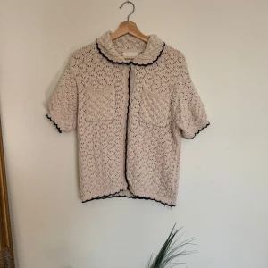 Beige stickad kofta från Zara - Beige stickad kofta från Zara med kort ärm, två fickor framtill och svarta vågiga detaljer längs kanterna och kragen. Koftan har ett luftigt, virkat mönster och stängs med knappar. Perfekt för lager-på-lager.