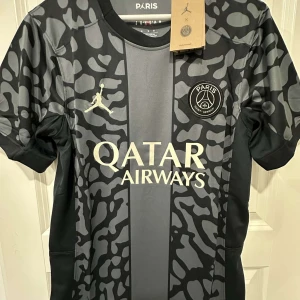 PSG x Jordan svart/grå fotbollströja - Snygg PSG x Jordan fotbollströja i svart och grått med unikt mönster och stora loggor på bröstet. Tröjan har korta ärmar, rund hals och är tillverkad i ett lätt, ventilerande material som passar perfekt för träning eller match.