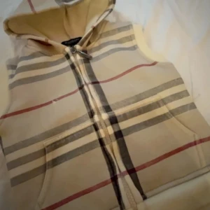 Rutig väst med huva från Burberry - Snygg väst från Burberry med klassiskt rutigt mönster i beige, grått, svart och rött. Väst med huva, dragkedja framtill och två fickor. Tillverkad i mjukt bomullsmaterial, perfekt för lager-på-lager-stil.