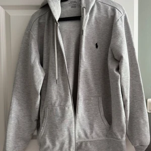 Grå hoodie från Polo Ralph Lauren - Snygg ljusgrå hoodie från Polo Ralph Lauren med dragkedja och klassisk broderad logga på bröstet. Tröjan har huva med snörning, två fickor fram och är tillverkad i mjuk bomullsmix. Perfekt för en chill och stilren look.