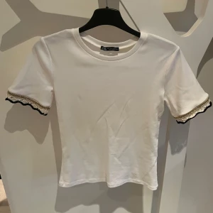 Zara tshirt med spets - Snygg vit topp från Zara med korta ärmar och dekorativa spetsdetaljer i svart och guld vid ärmsluten. Helt oanvänd och är slutsåld på hemsidan❣️