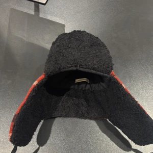 Rutig trappermössa från Nudie Jeans - Säljer en röd och svart rutig trappermössa från Nudie Jeans. Utsidan är i ullblandning med klassiskt rutmönster och insidan är fodrad med mjuk svart teddy. Mössan har öronlappar och knytband för extra värme och stil.