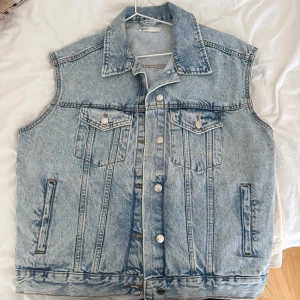 Ljusblå jeansväst med knappar - Säljer en klassisk ljusblå jeansväst med krage och silverfärgade knappar framtill. Västen har två bröstfickor med lock och knapp samt två sidofickor. Perfekt att slänga över en hoodie eller t-shirt för en cool look.