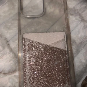 Glittrig mobilskal med plånboksficka i rosa  - Ett mobilskal till iPhone 13 Pro Max. Tänkte sälja för 50kr men såg att lite av kortfickans glitter hade åkt av så säljer för 25kr