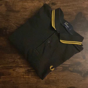 Fred Perry Pikétröja  - Fred Perry piké i svart med gula detaljer, storlek M. Ikonisk modell som passar perfekt för casual-stilen. Välanvänd med små hål och slitage, men fortfarande snygg med rätt attityd. Perfekt för dig som gillar vintage vibe och klassisk streetwear. Pris kan diskuteras pga slitage.