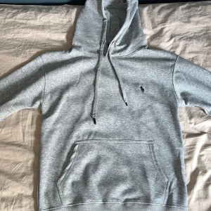 Grå Ralph Lauren hoodie - Snygg grå hoodie från Ralph Lauren med klassisk broderad logga på bröstet. Tröjan har huva med snörning, känguruficka och ribbade muddar. Perfekt för chill dagar och enkel att matcha med det mesta. Materialet är mjuk bomullsmix som känns skön mot huden. Ingen retur.