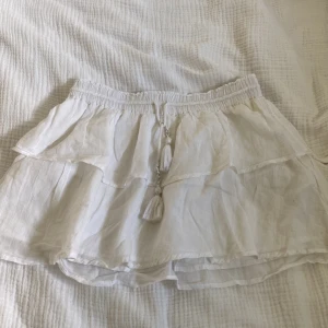 Vit volangkjol med tofsdetaljer - Superfin vit kortkjol i luftigt tyg med volanger och snygga tofsar med pärlor i midjan. Perfekt för varma dagar och ger en bohemisk vibe. Resår i midjan för skön passform.