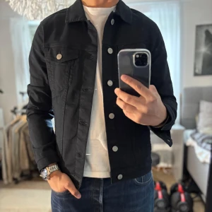 Svart jeansjacka Jack & Jones S - Stilren svart jeansjacka från Jack & Jones i klassisk modell med silverfärgade knappar. Jackan har två bröstfickor med lock och knapp, samt raka sidfickor. Perfekt för dig som gillar en clean och tidlös look.