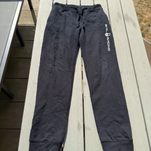 Svarta mjukisbyxor Sail Racing - Svarta mjukisbyxor från Sail Racing med logga på ena benet. Byxorna har snörning i midjan, muddar vid bensluten och en bakficka. Sköna och perfekta för chill eller träning. Tillverkade i mjukt bomullsmaterial.