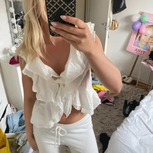 Vit volangblus från H&M - Supersöt vit blus från H&M med volanger och knyt framtill. Den är ärmlös och har en luftig, flowy passform som känns riktigt somrig. Perfekt för dig som gillar romantiska detaljer och en lätt, fräsch look.