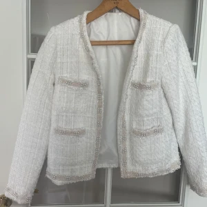 Vit kavaj med pärldetaljer - Snygg vit kavaj i tweed-look med pärldetaljer längs kanter och fickor. Jackan har fyra fickor framtill och är helfodrad. Perfekt för dig som vill ha en chic och elegant stil med lite extra bling.