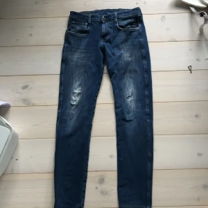 Blå slim jeans med slitningar - Säljer ett par blå slim jeans med snygga slitningar och tvättade detaljer på benen. Jeansen har klassisk femficksdesign, knappgylf och raka bakfickor med dekorativa sömmar. Perfekta för en avslappnad och trendig look.pris kan diskuteras.bred 28 men man kan ha den om man har upp till 30 i bredd för att det har anbass som marterial som är lite mer att man kan stretcha ut den 