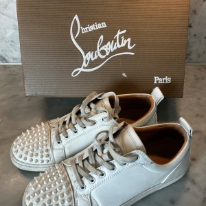 Vita sneakers från Christian Louboutin - Snygga vita sneakers från Christian Louboutin med ikoniska röda sulor och coola nitar på tån. Skorna har snörning och är tillverkade i skinn för en lyxig känsla. Skriv om minsta lila fundering !! Vet ej om äkta