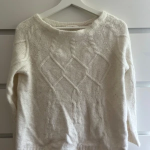 Vit stickad tröja Holly & Whyte - Mysig vit stickad tröja från Holly & Whyte med flätmönster framtill. Tröjan är ganska tjock och långa ärmar, perfekt för kyliga sommar dagar. Den är mjuk och har en klassisk, avslappnad passform som funkar till allt.