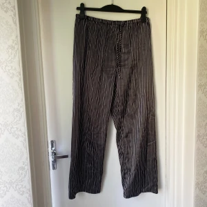 Randiga svarta pyjamasbyxor från Body Zone - Svarta byxor med vita vertikala ränder från Body Zone. De har en avslappnad passform, resår i midjan och snörning för extra komfort. Tillverkade i mjukt bomullsmaterial, perfekta för chill dagar eller som loungewear.