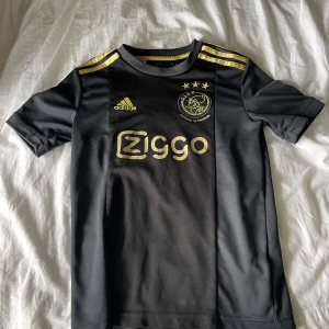 Ajax svart fotbollströja Adidas guld - En limited edition Ajax fotbollströja som inte längre går att få tag i. Köpt i Amsterdam i deras shop! Guldiga detaljer med en lite mörkare svart detalj i mitten av tröjan. Ziggo är en sponsor som Ajax har och det står stort på bröstet av tröjan.