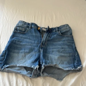 Blå jeansshorts från Lindex - Snygga blå jeansshorts från Lindex med råa kanter och klassisk femficksdesign. Shortsen har bälteshällor, knapp och dragkedja framtill samt bakfickor. Perfekta för sommaren och ger en avslappnad vibe.