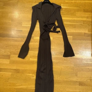 Brun ribbad långklänning  - Snygg brun långklänning från gina med ribbad struktur, långa ärmar och krage. Klänningen har öppning vid brösten med knyt  och utsvängda ärmslut. Perfekt för dig som gillar en stilren och trendig look.