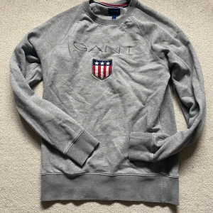 Grå sweatshirt från GANT med brodyr - Snygg grå sweatshirt från GANT med broderad logga och sköld på bröstet. Tröjan har rund halsringning, långa ärmar och ribbade muddar. Perfekt för en avslappnad och stilren look.
