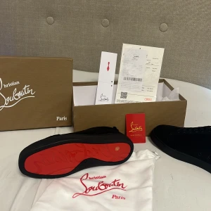 Christian Louboutin svarta sneakers - Exklusiva svarta sneakers från Christian Louboutin med ikonisk röd sula och diskret logga. Skorna har rund tå, snörning och är tillverkade i lyxigt skinn. Perfekta för dig som vill sticka ut med en stilren och lyxig vibe.