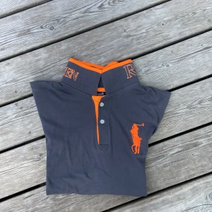 Mörkgrå pikétröja från Ralph Lauren - Snygg mörkgrå pikétröja från Ralph Lauren med orange detaljer och stor broderad logga på bröstet. Tröjan har korta ärmar, klassisk krage och orange kantband samt nummerdetalj på ärmen. Perfekt för en sportig och stilren look. Priset är ej hugget i sten och kan ändras.