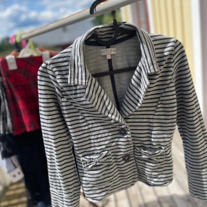 Randig kavaj från Indigo Collection - Snygg, söt grå och mörkblå randig kavaj från Indigo Collection. Kavajen har två framfickor med rosett-detaljer och knäpps med två knappar framtill. Mjuk bomullskänsla och klassisk krage, perfekt för en chill och stilren look.