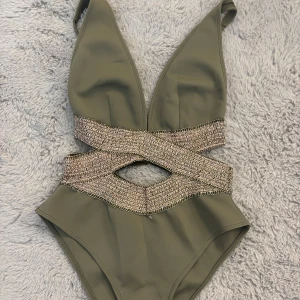 Olivgrön baddräkt med glittriga detaljer - Supersnygg olivgrön baddräkt från Luli Fama med djup v-ringning och glittriga band i midjan som korsas framtill. Utskuren detalj vid magen och justerbara axelband. Perfekt för stranden eller poolen när du vill sticka ut.
