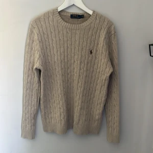 Beige kabelstickad tröja Polo Ralph Lauren - Kabelstickad beige tröja från Polo Ralph Lauren med klassisk rund hals och broderad logga på bröstet. Långärmad och ribbade muddar för en snygg passform. Perfekt för dig som gillar stilren och tidlös design.