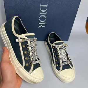 Dior sneakers dam - Snygga sneakers från Dior i marinblå canvas med vita detaljer och robust vit sula. Skorna har unika snören med texten 'Christian Dior J'Adior' och guldfärgade öljetter. Perfekt för dig som vill sticka ut med exklusiv streetstyle.
