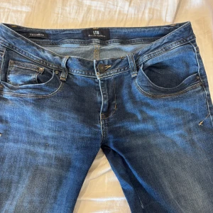 Blå jeans från LTB Valerie W32/L36 - Snygga blå jeans från LTB, modell Valerie, med klassisk femficksdesign och kontrastsömmar. Jeansen har normal passform och raka ben, tillverkade i mjukt denimtyg. Perfekta för dig som gillar en tidlös look med lite slitningar och cool tvätt.
